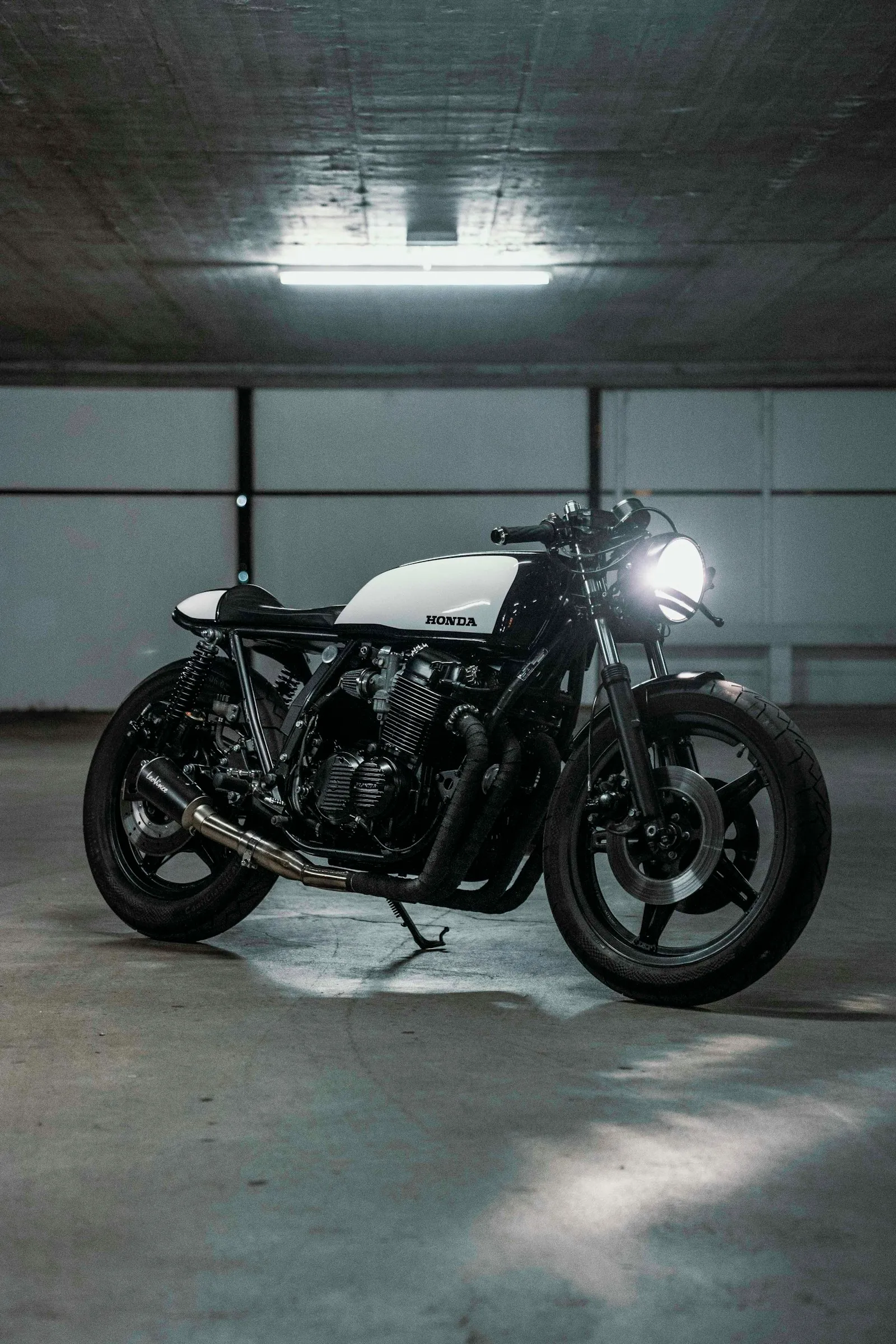 A black and white Honda CB cafe racer under a single striplight in a bare parking garage