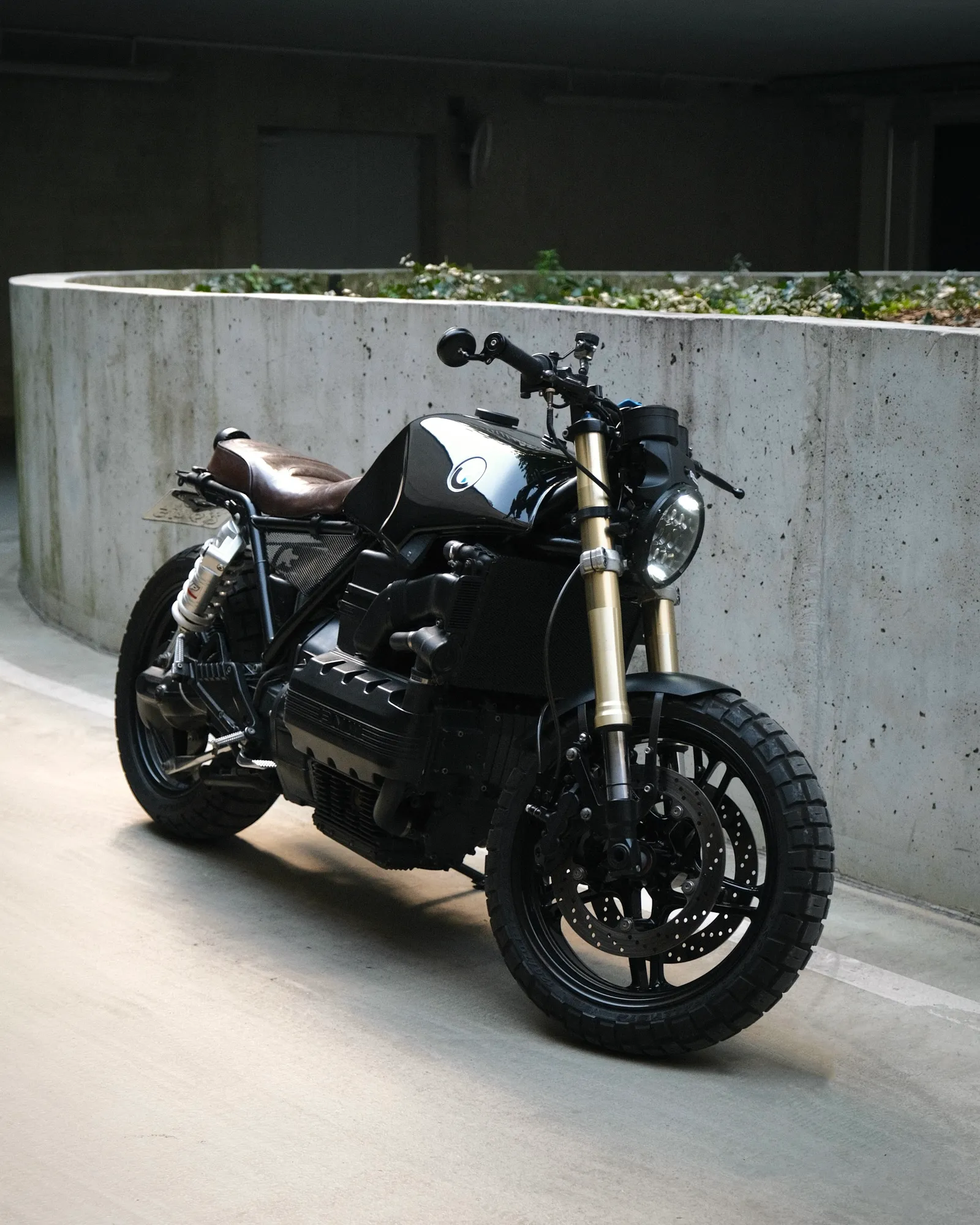 Front three-quarter view of the finished black K1100 with gold R1 forks and knobby Scorpion Rally tyres against a concrete wall